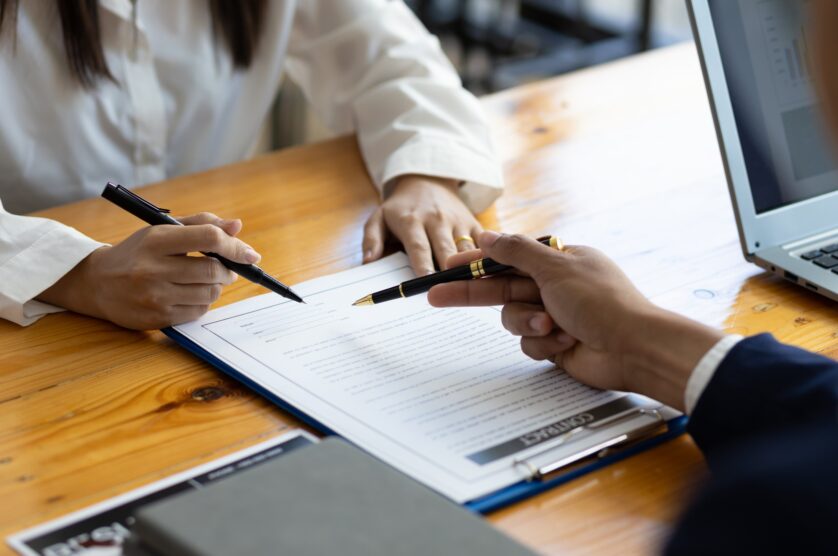 Two people are sitting at a wooden table, reviewing and discussing a document with pens in hand. A laptop is partially visible on the right side.