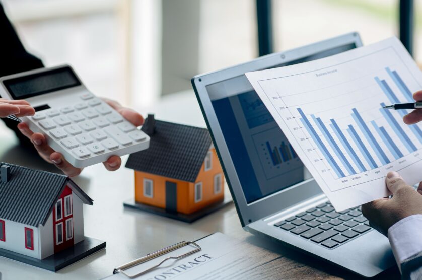 Two people discussing financial data. One holds a calculator, the other a paper with a bar chart. Small house models and a contract on the table.